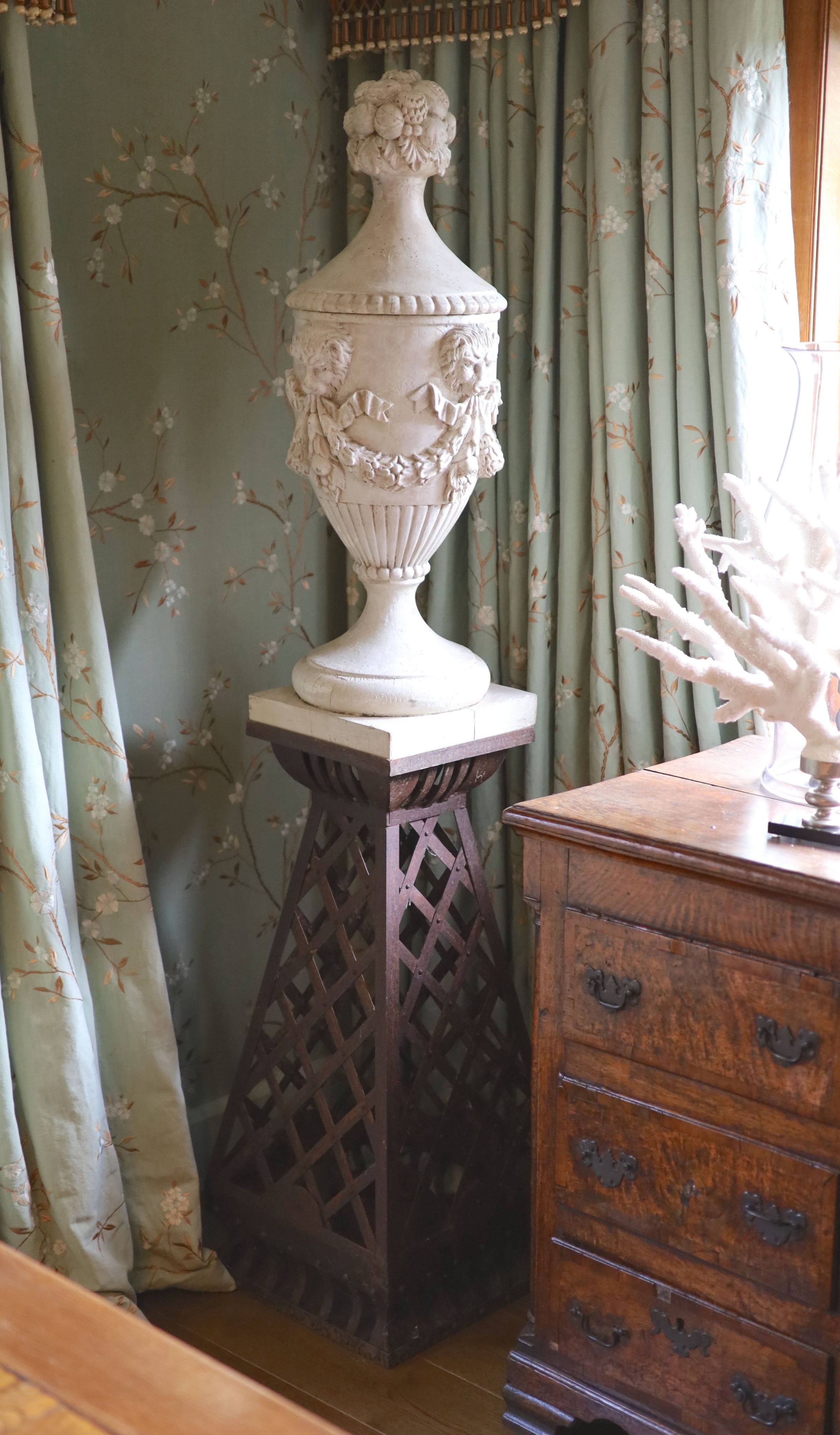 A pair of white painted carved walnut urns and covers, on wrought iron stands, the urns carved with fruit, lions masks and floral swags, height 90cm, the tapered stands with lattice work sides, height 90cm, overall heigh